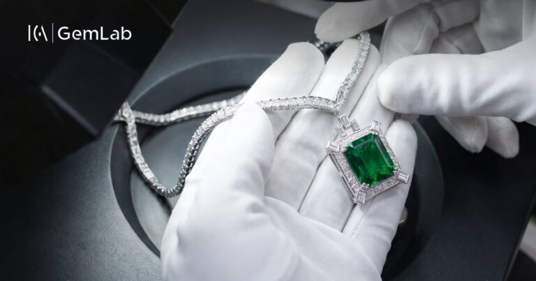 An emerald and diamond pendant held in gloved hands at ICA | GemLab