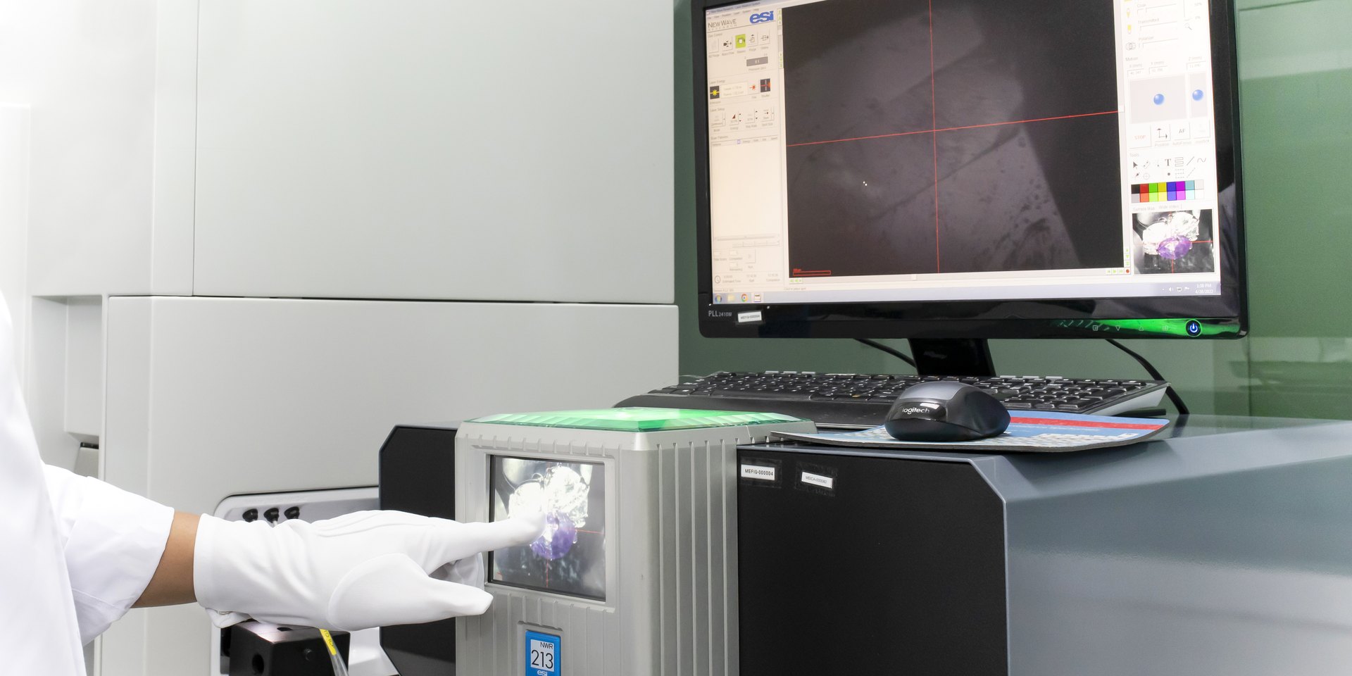 A gloved hand at the laser ablation cell of the LA-ICP-MS, with the targeting view of a stone on the screen behind.