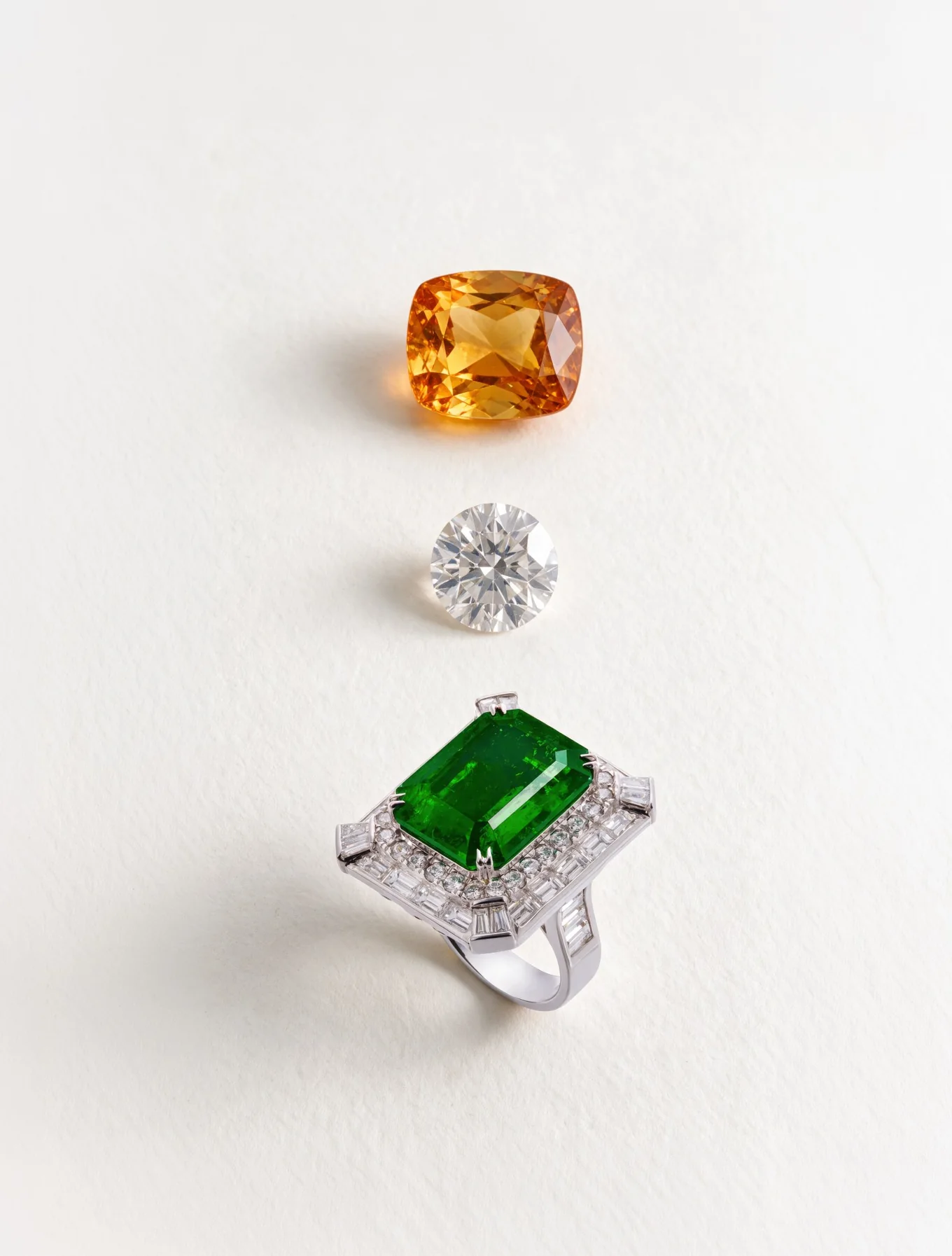 An orange cushion-cut gemstone, a round brilliant diamond and an emerald and diamond ring, arranged in a column on a pale ground.