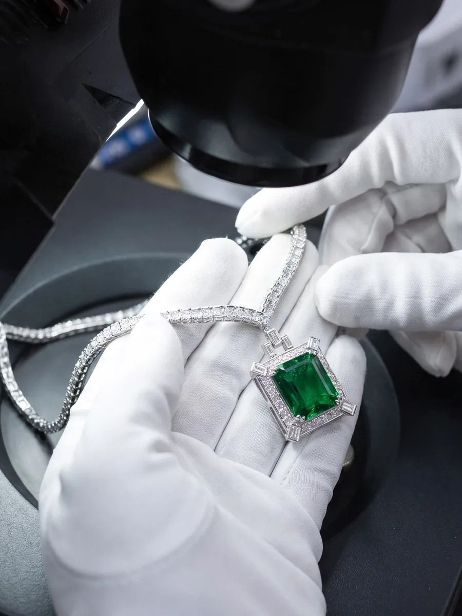 Illustration of gloved hands holding an emerald and diamond pendant beside a microscope.